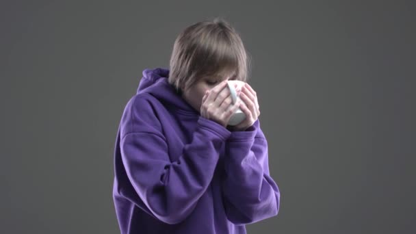 Caucasiano millennial jovem bebendo chá quente se aquece abraçando xícara no estúdio, vídeo 4k em fundo cinza — Vídeo de Stock