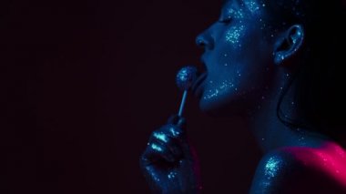 Mavi ışıkta parıldayan, lolipop yalayan kadın manken, ışıltılı vücut sanatıyla seksi kız, videoyu kapat.