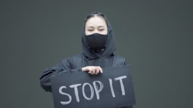 Siyah maskeli, elinde karton levha olan öfkeli kızı protesto ediyor. Gri stüdyo arka planında durduruyor.