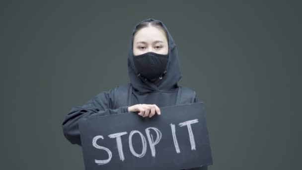 抗议戴着黑色面具拿着印有题词的硬纸板标语的愤怒女孩，在灰色工作室的背景下阻止了它 — 图库视频影像