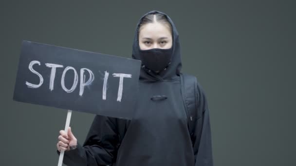 En protest mot grymhet och rasism. ung flicka i svart mask med skylt stoppa det i studio på grå bakgrund — Stockvideo