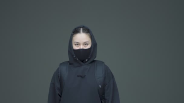 Νεαρή φεμινίστρια ακτιβίστρια με μαύρη κουκούλα και μάσκα πάει και σταματά στο γκρίζο φόντο του στούντιο — Αρχείο Βίντεο
