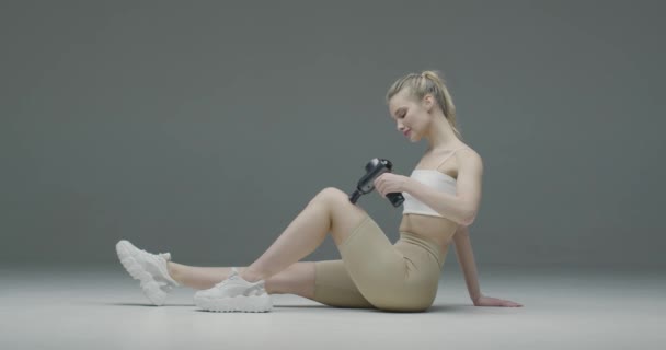 Atletyczna młoda blondynka masuje nogę przez ręczny masażer perkusyjny na szarym tle studio dla ulgi mięśni — Wideo stockowe
