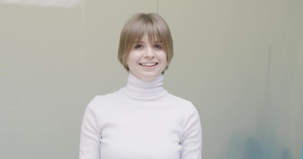 Позитивно дружелюбная улыбающаяся юная кавказская девушка в белой водолазке смотрит на камеру, позирующую на сером фоне студии — стоковое видео