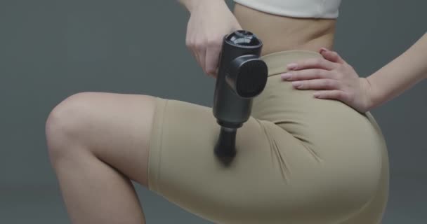 Молодая спортивная девушка в спортивной одежде массирует бедро с помощью массажного пистолета на сером фоне студии, перед тренировкой разминки, крупным планом — стоковое видео