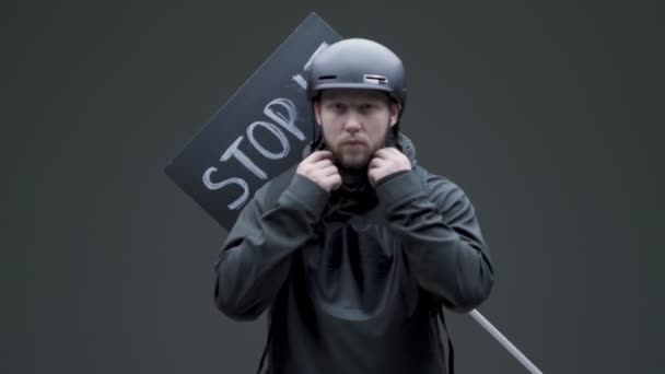 Мужчина-активист в черной одежде и шлеме надевает маску, готовится к пикету на сером фоне студии — стоковое видео