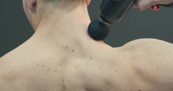 Масажер глибоких тканин, спортивний хлопець масажує шию для полегшення болю на сірому фоні студії, вид ззаду — стокове відео