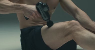 Atletik erkek sporcu el masajı tabancasıyla kalça masajı, egzersiz sonrası iyileşme rutinleri, yakın çekim.