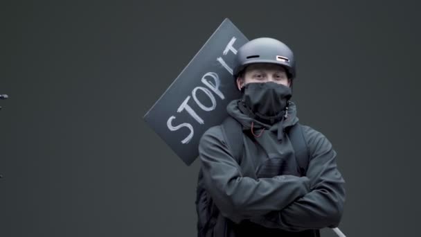 Mężczyzna protestujący w czarnej masce i kask z kartonowym znakiem stop w pozycji obronnej na szarym tle studio — Wideo stockowe