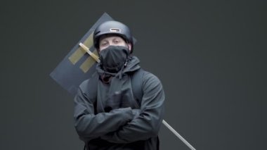 Siyah elbiseli, miğferli, maskeli protestocu. Protesto işaretli. Poz veriyor ve gri arka planda grev yapıyor.