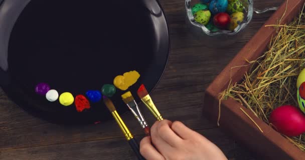 Pinceles en paleta. Vista superior de cerca el vídeo de la mano femenina con pinceles y pinturas durante la pintura en huevos de Pascua — Vídeo de stock