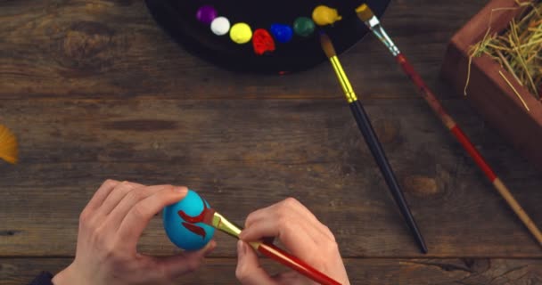 Top widok artystów ręce malowanie na jajku wielkanocnym pędzlem na drewnianym miejscu pracy z paletą farb — Wideo stockowe