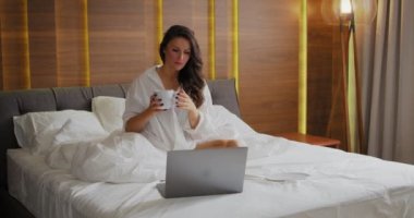 4K. Dizüstü bilgisayarlı güzel bir kadının yatak odasında beyaz yatakta çay içerken videosu.