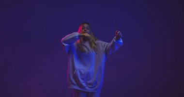 4K video, şık bir genç kız, neon ışıklı ve arka planda kapüşonlu bir kapüşonluyla dans ediyor..