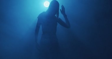 Modaya uygun genç bir kızın 4K videosu, gecenin karanlığında sisli mavi ışıkta dans etmek. Gizemli modern dans