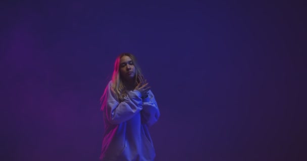 Stijlvol jong meisje, cool dansen in een hoodie in neon licht en gekleurde achtergrond in studio, kopieer ruimte — Stockvideo