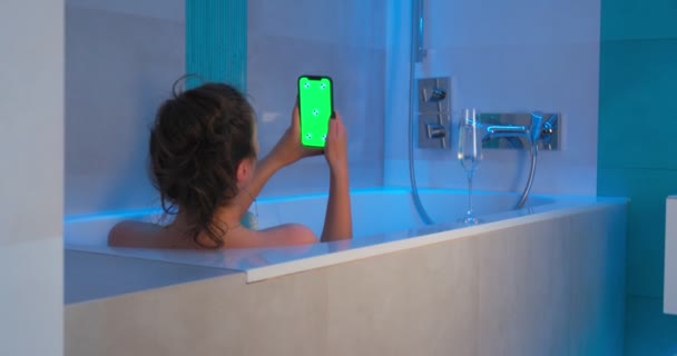 Samotna kobieta trzyma telefon z zielonym ekranem ang chromakey makieta w wannie. Dziewczyna oglądanie wideo leżące w łazience — Wideo stockowe