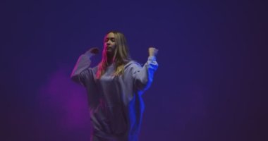 Neon renkli ışıkta kapüşonlu kapüşonlu kapüşonlu hip hop dansı yapan havalı genç kız. Çağdaş dansın 4k videosu
