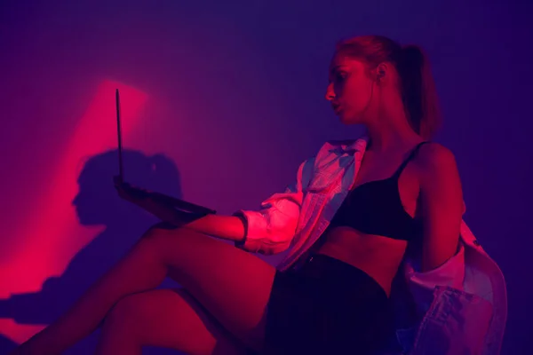 Молодая красивая девушка создает классный проект на ноутбуке в красочном неоновом свете. Мода модель с компьютером — стоковое фото