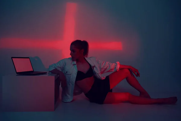 Креативная крутая молодая девушка с ноутбуком в красном студийном свете, сидящая на полу. Мода модель блоггер с компьютером. — стоковое фото