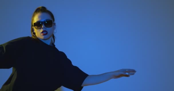 Menina de moda jovem elegante em óculos de sol dança legal em luz de estúdio de néon azul. Dança hip hop moderna. Espaço de cópia — Vídeo de Stock