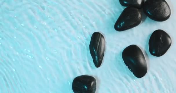 Fekete kövek fekszenek a kék vízben, amit a szél hullámzott. Tiszta víz texturált természetes háttér, felülnézet. Pihenési koncepció — Stock videók