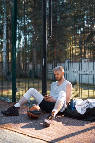 Basketbolcu havalı adam antrenmandan sonra dinleniyor. Yazın sahada telefonla oturuyor.