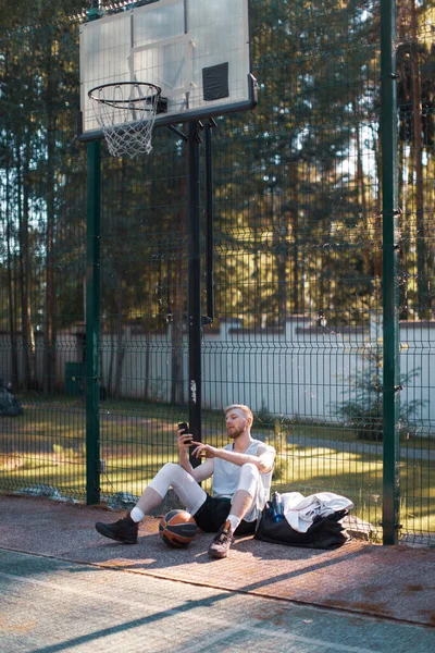 Баскетболист спортивный парень после тренировки сидит с телефоном на корте летом — стоковое фото