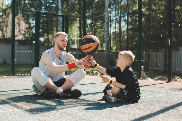 Instrutor de jogador de basquete profissional colocando bola no dedo de pequeno garoto esportivo sentado na quadra ao ar livre — Fotografia de Stock