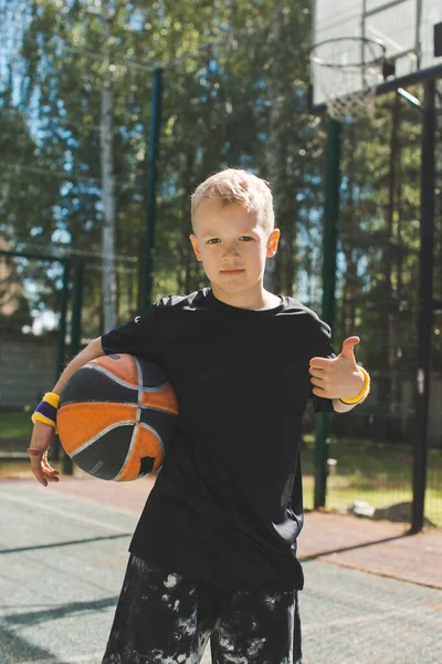 Elegante menino esportivo com bola de basquete mostra polegar acima gesto à luz do sol. Estilo de vida saudável das crianças — Fotografia de Stock