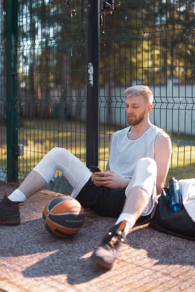 Baloncesto jugador fresco chico teniendo descanso después de entrenar sentado con el teléfono en la cancha al aire libre en verano — Foto de Stock