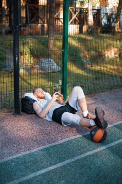 Basketbolcu sporcu adam basketbol sahasında antrenmandan sonra telefonla dinleniyor.