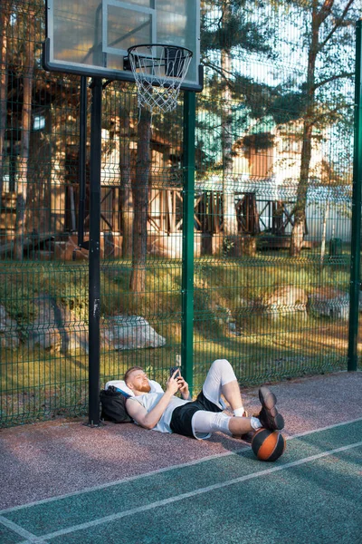 Чоловік баскетболіст лежить розслабляючись за допомогою додатка спортивного телефону після тренування на відкритому повітрі під баскетбольним кільцем — стокове фото