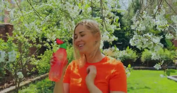 Souriant jeune belle femme buvant de la bouteille de sport dans le jardin en fleurs. Style de vie respectueux de l'environnement. Beauté naturelle — Video