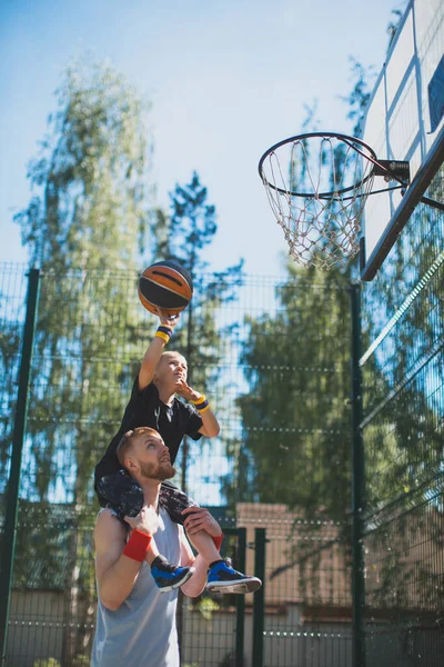 Счастливая семья, молодой спортивный отец, держащий сына на плечах. Мальчик бросает мяч в баскетбольное кольцо на открытом воздухе — стоковое фото