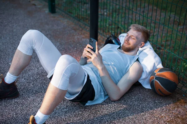 Баскетболист спортивный молодой парень лежит с телефоном с помощью социальных сетей приложений, имеющих перерыв на открытом воздухе на спортивной площадке — стоковое фото
