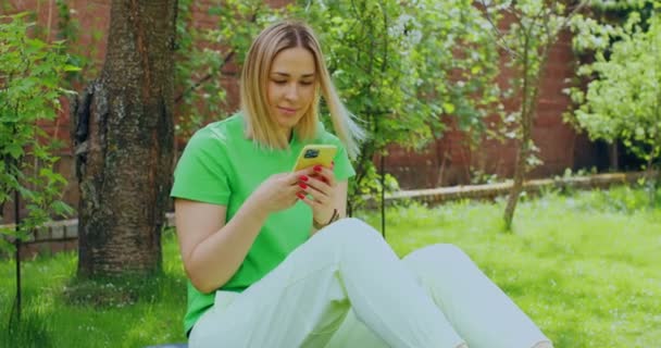 Όμορφη νεαρή γυναίκα blogger με τηλέφωνο κάθεται στο πράσινο γκαζόν στον κήπο, κουβεντιάζοντας στα κοινωνικά δίκτυα χρησιμοποιώντας εφαρμογές — Αρχείο Βίντεο