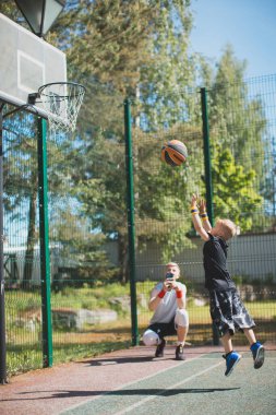 Küçük sporcu çocuk basketbol potasına top atıyor dışarıda antrenman yapıyor, baba kameraya video çekiyor.