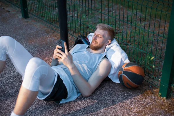 Крутой баскетболист спортивный молодой парень лежал расслабляясь с телефоном после тренировки на открытом воздухе на спортивной площадке — стоковое фото