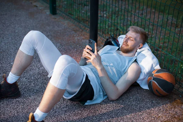 Баскетболист молодой спортсмен в спортивной одежде лежит с телефоном после тренировки на открытом воздухе на спортивном корте — стоковое фото