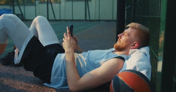スポーツコートで屋外でトレーニングした後、電話でリラックスして横たわってクールなバスケットボール選手のスポーティな若い男 — ストック動画