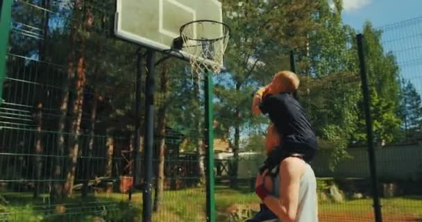 Jugadores de baloncesto. Joven padre deportivo sosteniendo al hijo sobre los hombros. Niño lanzando pelota en el aro de baloncesto al aire libre — Vídeos de Stock