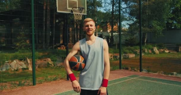 Jongeman met een sportbal op het basketbalveld glimlachend. Zelfverzekerde blanke mannelijke speler poseren outdoor in de zomer — Stockvideo