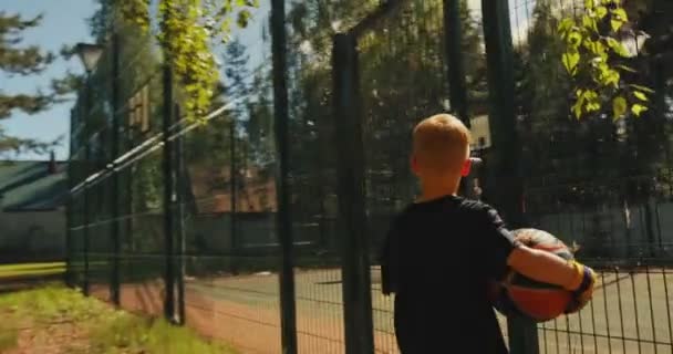 Jugador de baloncesto niño con pelota yendo a la cancha de baloncesto y anotando aro al aire libre en verano. Vista trasera — Vídeos de Stock