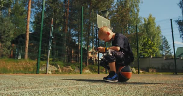 Jugador de baloncesto niño sentado en la pelota utilizando la aplicación de teléfono, viendo vídeo. Colegial jugando en línea en la cancha de baloncesto — Vídeos de Stock