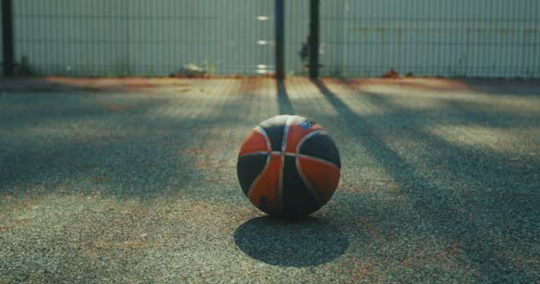 Primer plano con el foco en la pelota acostado en el suelo en la cancha de baloncesto al aire libre. Hombre recogiendo la pelota, luego alejándose — Vídeos de Stock