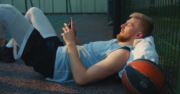 Cool basketbalspeler sportieve jonge kerel liggen ontspannen met de telefoon na het sporten buiten op het sportveld — Stockvideo