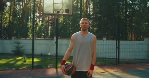 Δροσερό αρσενικό μπασκετμπολίστας ποζάρουν με μπάλα, σκουπίστε ιδρώτα από το μέτωπο στο γήπεδο μπάσκετ στο φως του ήλιου — Αρχείο Βίντεο