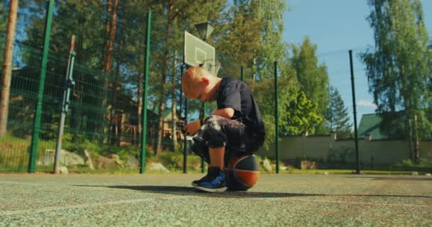 バスケットボール選手の少年は、電話アプリを使用してボールに座って、ソーシャルネットワークでコンテンツを見て、裁判所でオンラインでプレイ — ストック動画