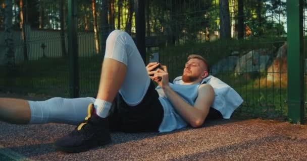 男性バスケットボール選手若いです男横たわっリラックス使用して電話アプリ後ワークアウト屋外 — ストック動画